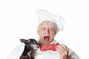80ish senior female in chef outfit is making faces while fighting a little dog for a piece of steak. Humor image.