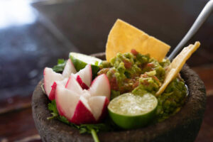 A high angle close up of chunky, delicious guacamole in a Mexican molcajete and crunchy corn chips, slices of lime and two radish halves, elegantly cut